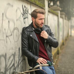 Fashionable man in a black leather jacket with headphones, leaning against a graffiti wall outdoors.